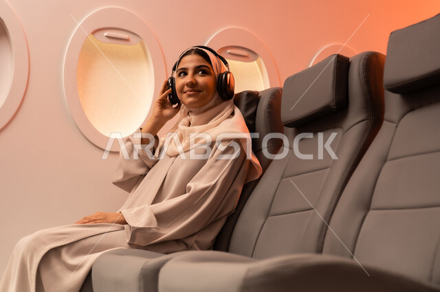 The concept of entertainment and amusement while riding an airplane, travel and tourism during the summer vacation, a close-up portrait from the side of a young Arab Gulf Omani woman wearing an abaya and hijab sitting on her seat inside the plane listening to music through headphones, preparing to spend an enjoyable tourist trip