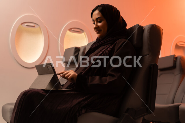Remote communication with family and friends, the concept of remote business management, entertainment and recreation, tourism during summer vacation, a close-up side portrait of a young Arab Gulf Omani woman working on an iPad while sitting on her seat inside the plane, Oman Air