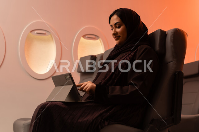 التواصل عن بعد مع الاهل و الأصدقاء، مفهوم اداره الاعمال والترفيه والتسليه عن بعد، السياحة في إجازة الصيف, بورتريه مقرب من الجانب لشابه عربيه خليجيه عمانيه تعمل على جهاز الايباد المحمول وهي تجلس على كرسيها داخل الطائره, الخطوط الجويه العمانيه