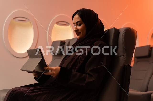 التواصل عن بعد مع الاهل و الأصدقاء، مفهوم اداره الاعمال والترفيه والتسليه عن بعد، السياحة في إجازة الصيف, بورتريه مقرب من الجانب لشابه عربيه خليجيه عمانيه تعمل على جهاز الايباد المحمول وهي تجلس على كرسيها داخل الطائره, الخطوط الجويه العمانيه