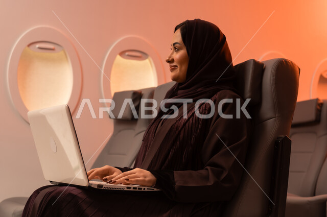 التواصل عن بعد مع الاهل و الأصدقاء، مفهوم اداره الاعمال والترفيه والتسليه عن بعد، السياحة في إجازة الصيف, بورتريه مقرب من الجانب لشابه عربيه خليجيه عمانيه تعمل على جهاز الايباد المحمول وهي تجلس على كرسيها داخل الطائره, الخطوط الجويه العمانيه