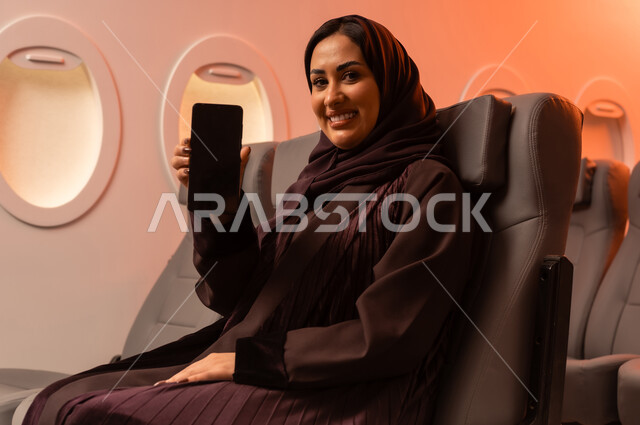 التواصل عن بعد مع الاهل و الأصدقاء، الإجابة والرد على الاتصالات باستخدام الهاتف المحمول، السياحة في إجازة الصيف, بورتريه مقرب من الجانب لشابه عربيه خليجيه عمانيه تمسك بهاتفهها المحمول على كرسيها داخل الطائره, الخطوط الجويه العمانيه