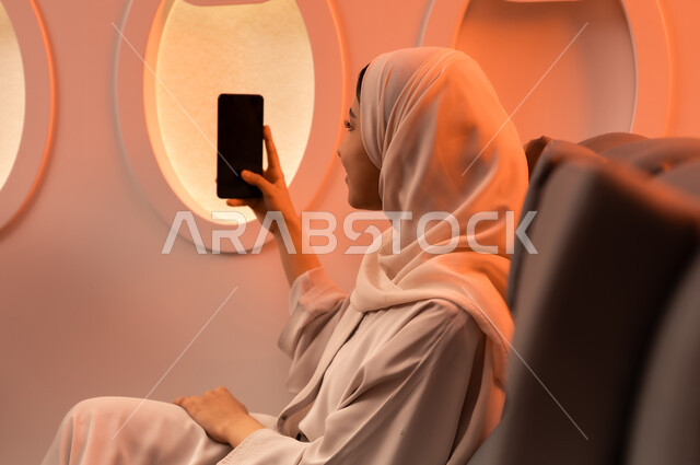 التواصل عن بعد مع الاهل و الأصدقاء، الإجابة والرد على الاتصالات باستخدام الهاتف المحمول، السياحة في إجازة الصيف, بورتريه مقرب من الجانب لشابه عربيه خليجيه عمانيه تمسك بهاتفهها المحمول على كرسيها داخل الطائره, الخطوط الجويه العمانيه