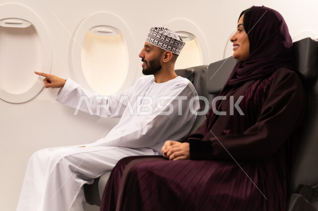 قضاء الاوقات السعيده ومفهوم التسليه والترفيه وتبادل الاحاديث داخل الطائره، بورتريه مقرب من الجانب لرجل وامراه عربيان خليجيان عمانيين يرتديان الزي العماني يجلسان على كرسيهما في رحله جويه يتبادلون الاحاديث ويشيرالرجل الى مكان ما من نافذه الطائره