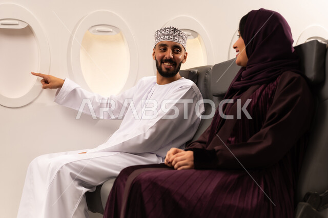 قضاء الاوقات السعيده ومفهوم التسليه والترفيه وتبادل الاحاديث داخل الطائره، بورتريه مقرب من الجانب لرجل وامراه عربيان خليجيان عمانيين يرتديان الزي العماني يجلسان على كرسيهما في رحله جويه يتبادلون الاحاديث ويشيرالرجل الى مكان ما من نافذه الطائره