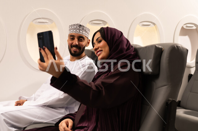 قضاء الاوقات السعيده داخل الطائره, مفهوم التسليه والترفيه وتبادل الاحاديث، بورتريه مقرب من الجانب لرجل وامراه عربيان خليجيان عمانيين يرتديان الزي العماني يجلسان على كرسيهما داخل الطائره في رحله جويه يقومون بالتقاط صور سيلفي