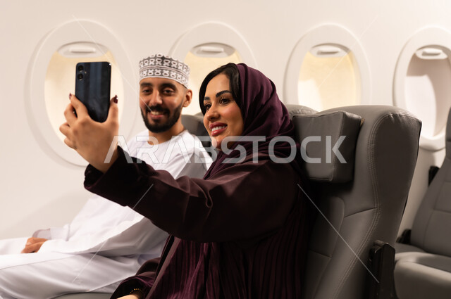 قضاء الاوقات السعيده داخل الطائره, مفهوم التسليه والترفيه وتبادل الاحاديث، بورتريه مقرب من الجانب لرجل وامراه عربيان خليجيان عمانيين يرتديان الزي العماني يجلسان على كرسيهما داخل الطائره في رحله جويه يقومون بالتقاط صور سيلفي
