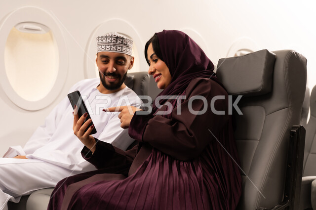 قضاء الاوقات السعيده داخل الطائره, مفهوم التسليه والترفيه وتبادل الاحاديث، بورتريه مقرب من الجانب لرجل وامراه عربيان خليجيان عمانيين يرتديان الزي العماني يجلسان على كرسيهما داخل الطائره في رحله جويه يقومون بالتقاط صور سيلفي