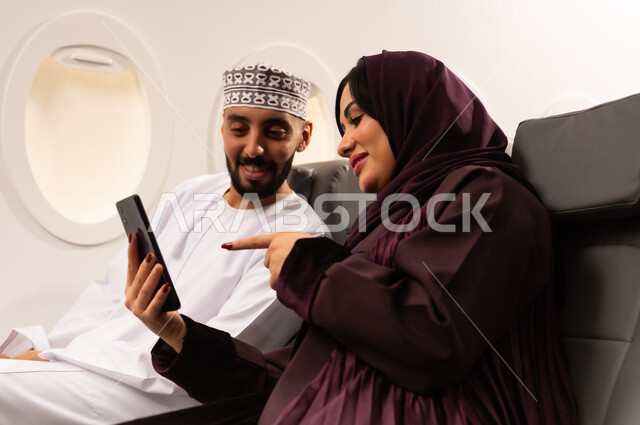 Having a good time inside the plane, the concept of entertainment, fun and chatting, a close-up side portrait of an Omani Gulf Arab man and woman wearing Omani dress sitting on their seats inside the plane during a flight, taking selfies