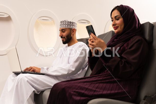 قضاء الاوقات السعيده داخل الطائره, مفهوم اداره الاعمال عن بعد، بورتريه مقرب من الجانب لرجل وامراه عربيان خليجيان عمانيين يرتديان الزي العماني يجلسان على كرسيهما داخل الطائره في رحله جويه يقوم الرجل بالعمل على جهاز الحاسوب المحمول وزوجته تتصفح هاتفهها