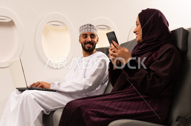 قضاء الاوقات السعيده داخل الطائره, مفهوم اداره الاعمال عن بعد، بورتريه مقرب من الجانب لرجل وامراه عربيان خليجيان عمانيين يرتديان الزي العماني يجلسان على كرسيهما داخل الطائره في رحله جويه يقوم الرجل بالعمل على جهاز الحاسوب المحمول وزوجته تتصفح هاتفهها