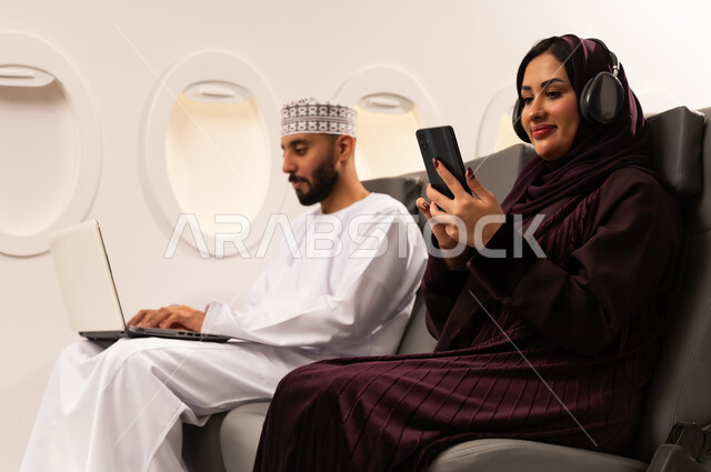 قضاء الاوقات السعيده داخل الطائره, مفهوم اداره الاعمال عن بعد، بورتريه مقرب من الجانب لرجل وامراه عربيان خليجيان عمانيين يرتديان الزي العماني يجلسان على كرسيهما داخل الطائره في رحله جويه يقوم الرجل بالعمل على جهاز الحاسوب المحمول وزوجته تقوم بالاستماع للموسيقى