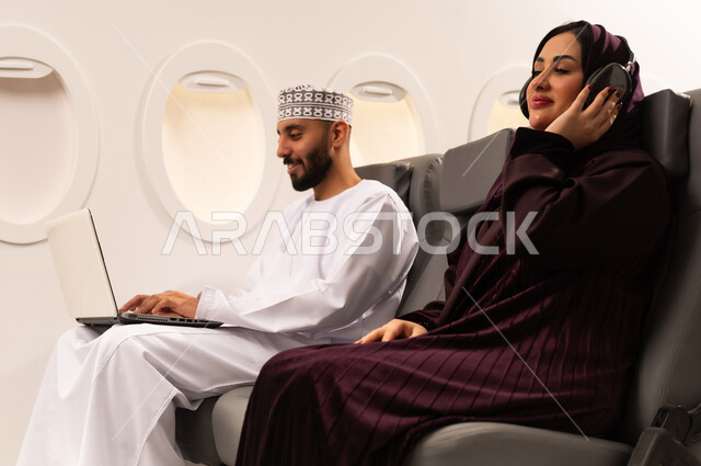 قضاء الاوقات السعيده داخل الطائره, مفهوم اداره الاعمال عن بعد، بورتريه مقرب من الجانب لرجل وامراه عربيان خليجيان عمانيين يرتديان الزي العماني يجلسان على كرسيهما داخل الطائره في رحله جويه يقوم الرجل بالعمل على جهاز الحاسوب المحمول وزوجته تقوم بالاستماع للموسيقى