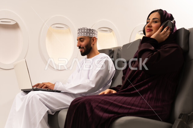قضاء الاوقات السعيده داخل الطائره, مفهوم اداره الاعمال عن بعد، بورتريه مقرب من الجانب لرجل وامراه عربيان خليجيان عمانيين يرتديان الزي العماني يجلسان على كرسيهما داخل الطائره في رحله جويه يقوم الرجل بالعمل على جهاز الحاسوب المحمول وزوجته تقوم بالاستماع للموسيقى