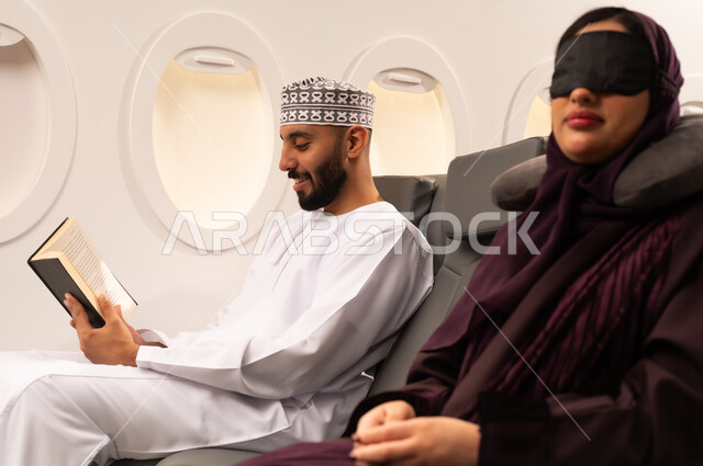 الترفيه والتسليه وقضاء الاوقات داخل الطائره, ممارسه الهوايات و الاستمتاع بقرائه القصص والروايات, بورتريه مقرب من الجانب لرجل وامرأه عربيان خليجيان عمانيين يرتديان الزي العماني يجلسان على كراسي الطائره في رحله جويه يقرأ الرجل كتابا وبجانبه زوجته النائمه