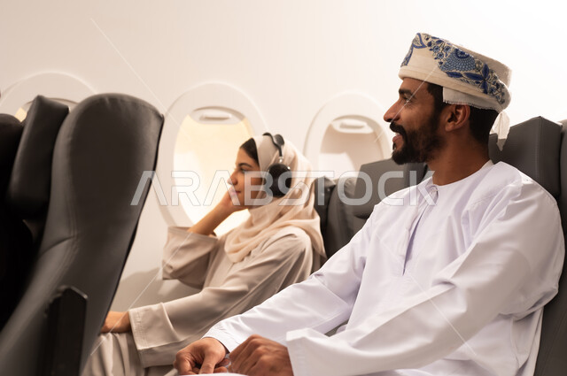 الترفيه والتسليه وقضاء الاوقات داخل الطائره, ممارسه الهوايات والاستماع الى الموسيقى, بورتريه مقرب من الجانب لرجل وامرأه عربيان خليجيان عمانيين يرتديان الزي العماني يجلسان على كراسي الطائره في رحله جويه يضعان سماعات الاذن (الهيدفوون)