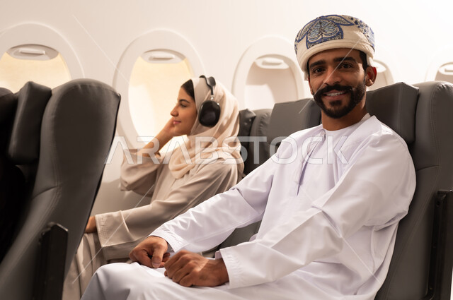 الترفيه والتسليه وقضاء الاوقات داخل الطائره, ممارسه الهوايات والاستماع الى الموسيقى, بورتريه مقرب من الجانب لرجل وامرأه عربيان خليجيان عمانيين يرتديان الزي العماني يجلسان على كراسي الطائره في رحله جويه يضعان سماعات الاذن (الهيدفوون)