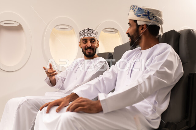 قضاء الاوقات السعيده داخل الطائره, التسليه والترفيه والاندماج مع الاصدقاء، بورتريه مقرب من الجانب لرجلان عربيان خليجيان عمانيين يرتديان الزي العماني يجلسان على كرسيهما داخل الطائره في رحله جويه يقومان بتبادل الاحاديث