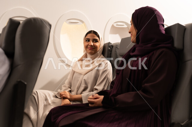 قضاء الاوقات السعيده داخل الطائره, التسليه والترفيه والاندماج مع الاصدقاء، بورتريه مقرب من الجانب لسيدتان عربيتان خليجيتان عمانيتين ترتديان العبائه والحجاب تجلسان على كرسيهما داخل الطائره في رحله جويه تقومان بتبادل الاحاديث
