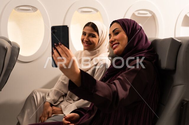 قضاء الاوقات السعيده داخل الطائره, التسليه والترفيه والاندماج مع الاصدقاء، بورتريه مقرب من الجانب لسيدتان عربيتان خليجيتان عمانيتين ترتديان العبائه والحجاب تجلسان على كرسيهما داخل الطائره في رحله جويه تقومان بالتقاط سيلفي بالهاتف المحمول