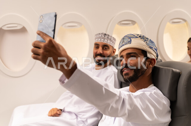 قضاء الاوقات السعيده داخل الطائره, التسليه والترفيه والاندماج مع الاصدقاء، بورتريه مقرب من الجانب لرجلان عربيان خليجيان عمانيين يرتديان الزي العماني يجلسان على كرسيهما داخل الطائره في رحله جويه يقومان بالتقاط سيلفي بالهاتف المحمول