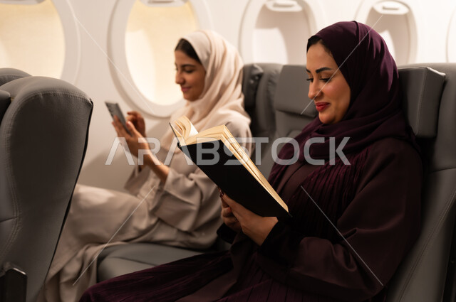 أداء المهام والواجبات العملية، الاندماج والتركيز لإنجاز المهام عن بعد، بورتريه مقرب من الجانب لسيدتان عربيتان عمانيتان ترتديان العبائه والحجاب تجلسان على كراسي الطائره في رحله جويه تقوم أحدهما بالعمل على جهاز الاب توب المحمول والاخرى تقرأ كتابا