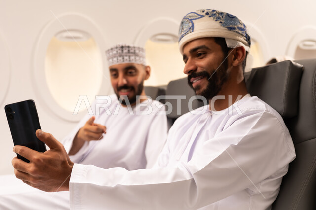 قضاء الاوقات السعيده داخل الطائره, التسليه والترفيه والاندماج مع الاصدقاء، بورتريه مقرب من الجانب لرجلان عربيان خليجيان عمانيين يرتديان الزي العماني يجلسان على كرسيهما داخل الطائره في رحله جويه يقومان بالتقاط سيلفي بالهاتف المحمول