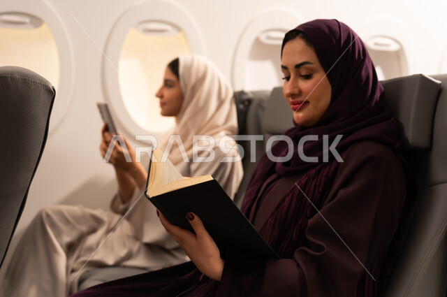 أداء المهام والواجبات العملية، الاندماج والتركيز لإنجاز المهام عن بعد، بورتريه مقرب من الجانب لسيدتان عربيتان عمانيتان ترتديان العبائه والحجاب تجلسان على كراسي الطائره في رحله جويه تقوم أحدهما بالعمل على جهازها المحمول والاخرى تقرأ كتابا