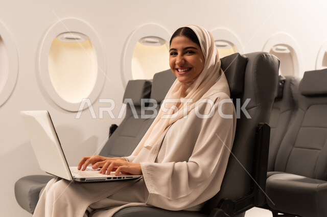 Performing practical tasks and duties, integration and concentration to accomplish tasks remotely, close-up side portrait of an Arab Gulf Omani woman wearing an abaya and hijab sitting on her seat inside an airplane on a flight working on a laptop