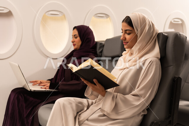 أداء المهام والواجبات العملية، الاندماج والتركيز لإنجاز المهام عن بعد، بورتريه مقرب من الجانب لسيدتان عربيتان عمانيتان ترتديان العبائه والحجاب تجلسان على كراسي الطائره في رحله جويه تقوم أحدهما بالعمل على جهاز الحاسب المحمول والاخرى تقرأ كتابا