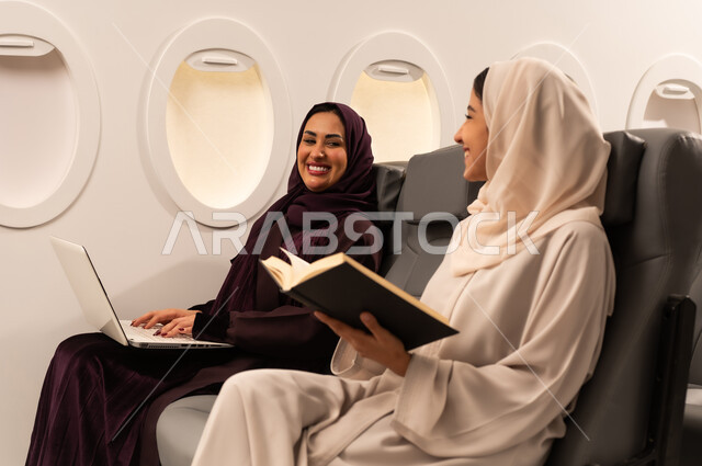أداء المهام والواجبات العملية، الاندماج والتركيز لإنجاز المهام عن بعد، بورتريه مقرب من الجانب لسيدتان عربيتان عمانيتان ترتديان العبائه والحجاب تجلسان على كراسي الطائره في رحله جويه تقوم أحدهما بالعمل على جهاز الحاسب المحمول والاخرى تقرأ كتابا