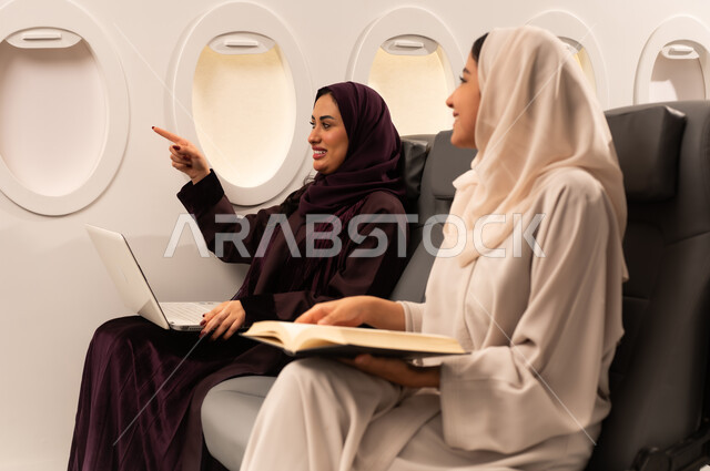 Performing practical tasks and duties, integration and concentration to complete tasks remotely, close-up side portrait of two Omani Arab women wearing abaya and hijab sitting on airplane seats on a flight, one of them is working on a laptop and the other is reading a book