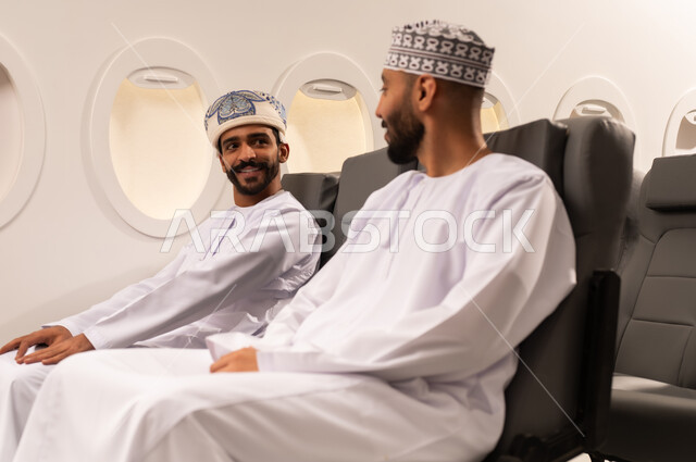 الاستمتاع بتبادل الاحاديث مع الاصدقاء, تعابير التسلية والمرح, بورتريه مقرب من الجانب لرجلان عربيان عمانيان خليجيان يرتديان الزي العماني يجلسون داخل مقاعد احدى الطائرات الخليجيه يتبادلون الاحاديث بسرور واهتمام, استغلال الاوقات في الدردشه