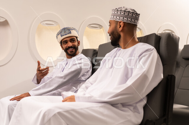 Enjoying chatting with friends, expressions of amusement and fun, a close-up side portrait of two Omani Gulf Arab men wearing Omani clothing sitting inside the seats of a Gulf airplane, exchanging conversations with pleasure and interest, taking advantage of the time to chat