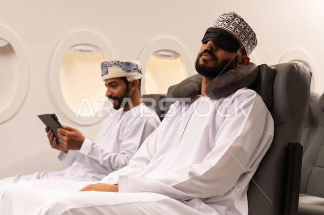 مفهوم السفر والسياحه وانجاز الاعمال داخل الطائره, بورتريه مقرب من الجانب لرجليين عربيين خليجيين عمانييان يرتديان الزي العماني يجلسان على مقاعدهما داخل احدى الطائرات الخليجيه يقوم احدهما بالنوم على مخده السفر ويغطي عينه بماسك اسود والاخر يستخدم الايباد