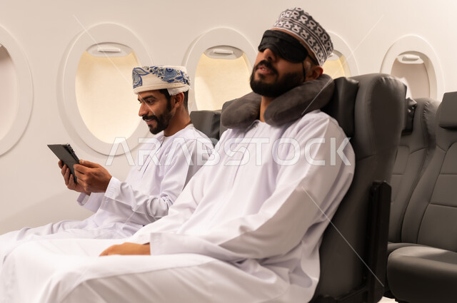 مفهوم السفر والسياحه وانجاز الاعمال داخل الطائره, بورتريه مقرب من الجانب لرجليين عربيين خليجيين عمانييان يرتديان الزي العماني يجلسان على مقاعدهما داخل احدى الطائرات الخليجيه يقوم احدهما بالنوم على مخده السفر ويغطي عينه بماسك اسود والاخر يستخدم الايباد