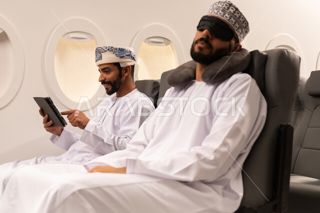 مفهوم السفر والسياحه وانجاز الاعمال داخل الطائره, بورتريه مقرب من الجانب لرجليين عربيين خليجيين عمانييان يرتديان الزي العماني يجلسان على مقاعدهما داخل احدى الطائرات الخليجيه يقوم احدهما بالنوم على مخده السفر ويغطي عينه بماسك اسود والاخر يستخدم الايباد