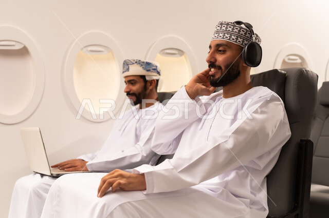 مفهوم السفر والسياحه وانجاز الاعمال داخل الطائره, بورتريه مقرب من الجانب لرجليين عربيين خليجيين عمانييان يجلسان على مقاعدهما داخل احدى الطائرات الخليجيه يرتدي احدهم سماعات الووكمان يستمع الى الموسيقى بسرور وسعاده والاخر يسستخدم جهازالاب توب المحمول