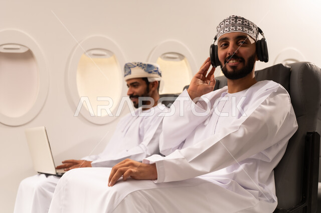 مفهوم السفر والسياحه وانجاز الاعمال داخل الطائره, بورتريه مقرب من الجانب لرجليين عربيين خليجيين عمانييان يجلسان على مقاعدهما داخل احدى الطائرات الخليجيه يرتدي احدهم سماعات الووكمان يستمع الى الموسيقى بسرور وسعاده والاخر يسستخدم جهازالاب توب المحمول