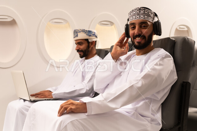 The concept of travel, tourism and completing business inside the plane, a close-up portrait from the side of two Omani Gulf Arab men sitting on their seats inside a Gulf plane, one of them wearing Walkman headphones, listening to music with pleasure and happiness, and the other using a laptop