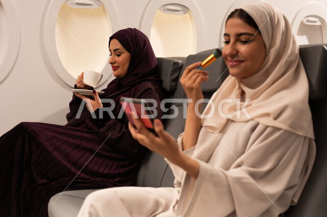 الاهتمام والعنايه بالمظهر الخارجي, التسويق لمستحضرات التجميل, بورتريه مقرب من الجانب لامرأتين عربيتين خليجيتين عمانيتيان ترتديان العباءة والحجاب تضع احداهما المكياج على وجهها داخل الطائره باستعمال فرشاة أحمر الخدود بينما تقوم الاخرى باحتساء كوبا من الشاي الساخن