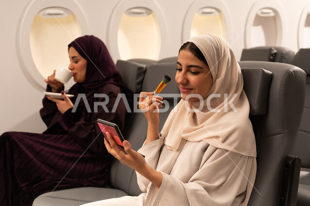 الاهتمام والعنايه بالمظهر الخارجي, التسويق لمستحضرات التجميل, بورتريه مقرب من الجانب لامرأتين عربيتين خليجيتين عمانيتيان ترتديان العباءة والحجاب تضع احداهما المكياج على وجهها داخل الطائره باستعمال فرشاة أحمر الخدود بينما تقوم الاخرى باحتساء كوبا من الشاي الساخن