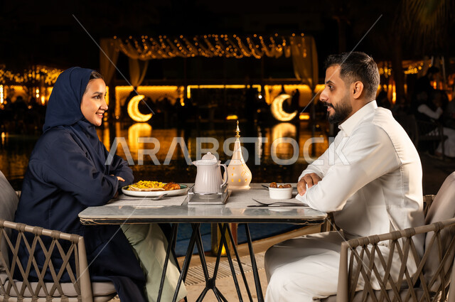 نظرات مليئة بالحب, مفهوم الرابطة الزوجية والألفة بين الزوجين, زوجان عربيان خليجيان سعوديان يجلسان في مطعم فاخر, الاستمتاع بالنقاشات والحوارات العائلية في الهواء الطلق,  روحنيات رمضانية, زينة وأنوار الأعياد والمناسبات