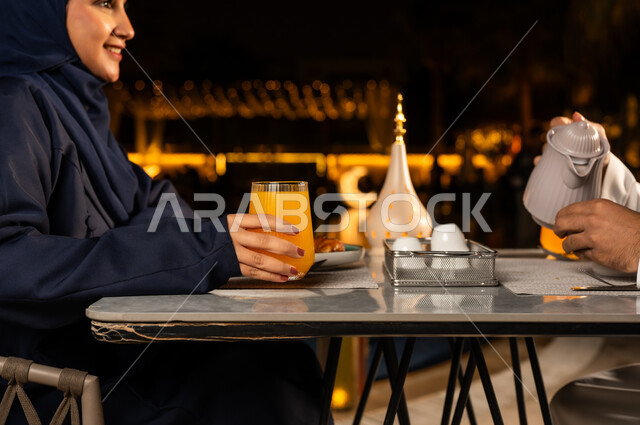 الاستمتاع بتناول المشروبات الطازجه, زينه و ديكور الشهر الكريم , روحنيات رمضانية ,سفرة مليئة باصناف من المأكولات اللذيذة ،صورة مقربة من الجانب لامراة عربيه خليجيه سعودية محجبه مبتسمه تقوم بامساك كأس من البرتقال بايماءات السعاده و السرور، فانوس رمضان