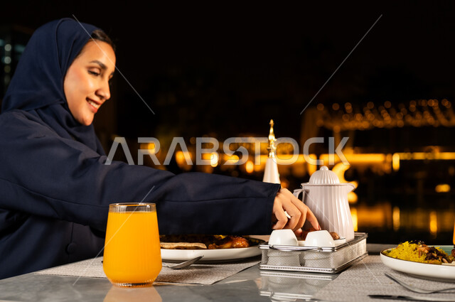 Gestures of happiness and joy, religious Ramadan atmosphere, close-up of a smiling Saudi Arabian Gulf woman wearing an abaya eating dates at the Maghrib call to prayer, a table full of delicious food, performing the ritual of fasting