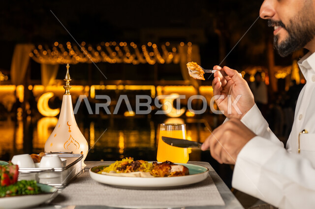 روحنيات رمضانية , زينه و ديكور الشهر الكريم ,سفرة مليئة باصناف من المأكولات اللذيذة ،صورة مقربة من الجانب ليد رجل عربي خليجي سعودي يرتدي الزي التقليدي يقوم بتناول الطعام على مائدة الافطار، ايماءات وجه سعيدة، فانوس رمضان