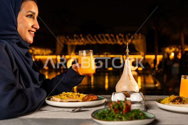 الاستمتاع بتناول المشروبات الطازجه, زينه و ديكور الشهر الكريم , روحنيات رمضانية ,سفرة مليئة باصناف من المأكولات اللذيذة ،صورة مقربة من الجانب لامراة عربيه خليجيه سعودية محجبه مبتسمه تقوم بامساك كأس من البرتقال بايماءات السعاده و السرور، فانوس رمضان
