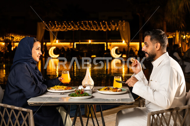 حوارات ونقاشات على مائدة الطعام, زينة وديكور شهر رمضان المبارك, قضاء وقت ممتع مع الأسرة, مائدة مليئة بالأطعمة والمشروبات الشهية، جلسة عائلية رمضانية روحانية, زوجان عربيان سعوديان خليجيان مبتسمان يرتديان الزي التقليدي يجلسان في مطعم, الاستمتاع الفطار