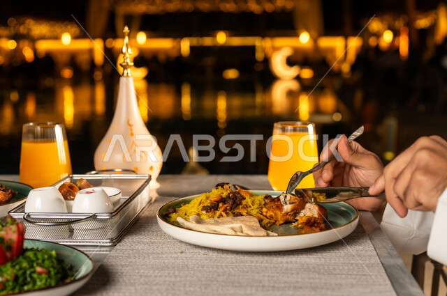 زينه و ديكور الشهر الكريم , روحنيات رمضانية ,سفرة مليئة باصناف من المأكولات اللذيذة ،صورة مقربة من الجانب ليد رجل عربي خليجي سعودي يرتدي الزي التقليدي يقوم بتناول الطبق الرئيسي على مائدة الافطار، فانوس رمضان, الاستمتاع بتناول المشروبات الطازجه