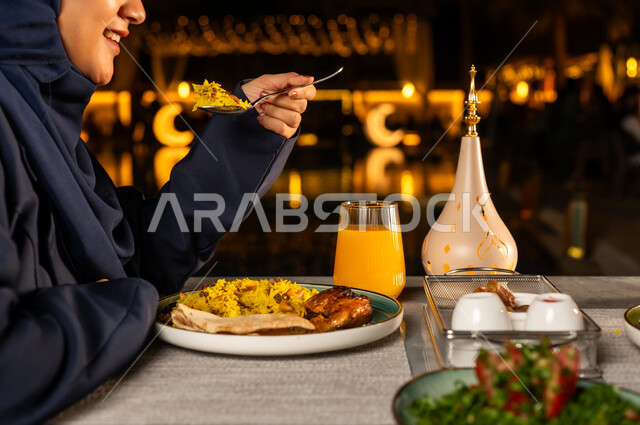 زينة شهر رمضان وسفرة مليئة باصناف من المأكولات اللذيذة وتناول وجبة الافطار ،صورة مقربة من الجانب ليد امراة عربيه خليجيه سعودية ترتدي العباءة تقوم بتناول الطعام على مائدة الافطار، ايماءات وجه سعيدة واجواء رمضانية، فانوس رمضان، شهر رمضان الكريم
